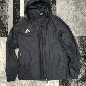 Svart vindjacka från Adidas - Säljer en svart vindjacka från Adidas med huva och dragkedja. Jackan har långa ärmar och två fickor. Perfekt för blåsiga dagar.
