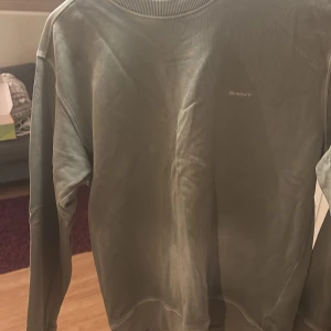 Grön tröja från GANT - Säljer en stilren grön tröja från GANT i storlek medium. Tröjan har en rund halsringning och ribbade muddar vid ärmslut och nederkant. Den är lite solblekt på magen och har en liten fläck vid loggan. Annars i bra skick