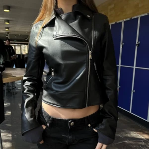 Svart skinnjacka från Kappahl - Snygg svart skinnjacka från Kappahl med asymmetrisk dragkedja och klassisk krage. Jackan har en croppad passform och långa ärmar. Perfekt för en cool och stilren look.