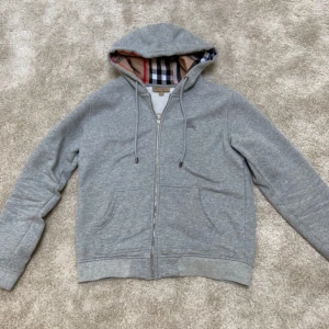 Grå hoodie från Burberry - Säljer en stilren grå hoodie från Burberry med dragkedja och klassiskt rutigt mönster i luvan. Om du är intresserad så skriv innan köp! 