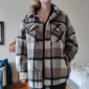 Rutig overshirt i beige och svart - Snygg och mysig jacka/overshirt i beige och svart med rutigt mönster, perfekt för vår/höst. Jackan har en oversized passform och är utrustad med knappar framtill. 