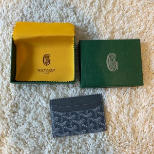 Grå plånbok  - Säljer en elegant svart plånbok från G🐡yard med deras ikoniska mönster. Plånboken är kompakt och perfekt för att hålla kort och kontanter. Den kommer i en snygg grön förpackning med gul insida.