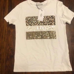 Vit t-shirt från Stradivarius - Säljer en vit t-shirt från Stradivarius med texten 'Nice PEOPLE' i guld. T-shirten har korta ärmar och detaljer i leopardmönster och paljetter. Perfekt för att ge din outfit lite extra glans!