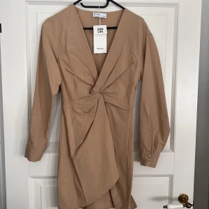 Beige klänning från Bershka ny med prislapp  - Snygg beige klänning från Bershka. Klänningen har långa ärmar och knappar vid ärmsluten. Perfekt för en stilren look.