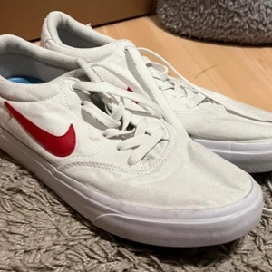 Vita sneakers från Nike - Snygga vita sneakers från Nike med röd swoosh på sidan. Skorna har en klassisk design med snörning och rund tå. Perfekta för en stilren look.