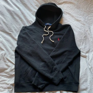 Svart hoodie från Ralph Lauren - Säljer en klassisk svart hoodie från Ralph Lauren med en liten röd logga på bröstet. Den har en justerbar huva med vita snören och en bekväm passform. Perfekt för en avslappnad stil. Mycket bra skick! Skriv vid minsta fundering. 