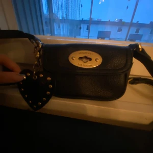 Svart axelväska från Mulberry - Snygg svart axelväska från Mulberry i skinn med ett hjärtformat hänge dekorerat med nitar. Väskan har en elegant metallplatta på framsidan och en justerbar axelrem. Perfekt för att bära dina viktigaste saker med stil. Dens knapp för att låsa är borta men den ser stängd ut ändå