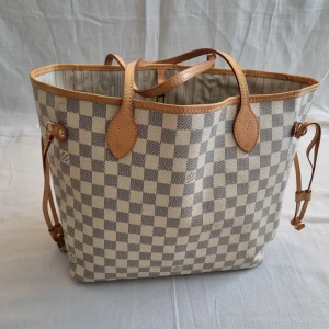 Rutig shoppingväska med läderdetaljer - damier  - Shoppingväska med klassiskt rutigt mönster i beige och grått. Väskan har läderhandtag och sidodetaljer i brunt läder. 