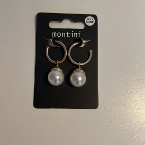 Guldörhängen med pärlor från Montini - Eleganta guldörhängen från Montini med stora vita pärlor som hänger från en halvcirkelformad design. Perfekta för att ge en stilren touch till din outfit.