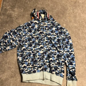 BAPE hoodie XXL (barn) - Säljer en cool hoodie från A Bathing Ape med blått och grått camouflage-mönster. Den har en unik design med hajmotiv på huvan och dragkedja framtill. Perfekt för streetwear-entusiaster! 