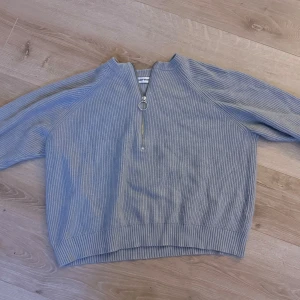 Grå ribbad tröja - Säljer en stilren grå ribbad tröja. Tröjan har en dragkedja framtill med en rund ringdetalj och långa ärmar. Perfekt för en avslappnad look. Passar bra till jeans eller kjol.