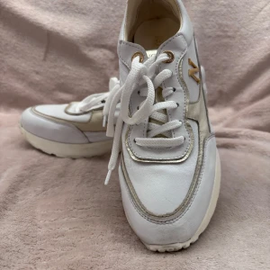 Vita sneakers med gulddetaljer - Snygga vita sneakers med gulddetaljer och snörning. Skorna har en tjock sula och en dekorativ guldfärgad häl. Perfekta för en stilren look.