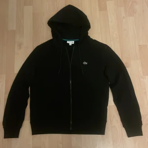 Svart hoodie från Lacoste - Säljer en stilren svart hoodie från Lacoste i 10/10 skick. Perfekt för en avslappnad look. Den har långa ärmar och en bekväm huva.