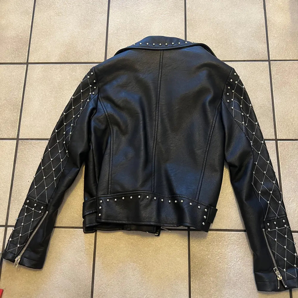 Snygg svart skinnjacka från Amisu med coola nitar och quiltade detaljer på ärmarna. Jackan har dragkedja och ett bälte i midjan för extra stil. Perfekt för att ge din outfit en edgy touch!. Takit.