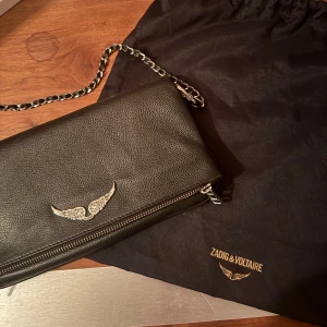 ÄKTA väska  från Zadig & Voltaire - Snygg svart skinnväska från Zadig & Voltaire med kedjeaxelrem och coolt vingmotiv. Väskan har en praktisk dragkedja framtill och är perfekt för att bära dina viktigaste saker. En stilren accessoar som passar till många tillfällen. De finns inga skador på väskan