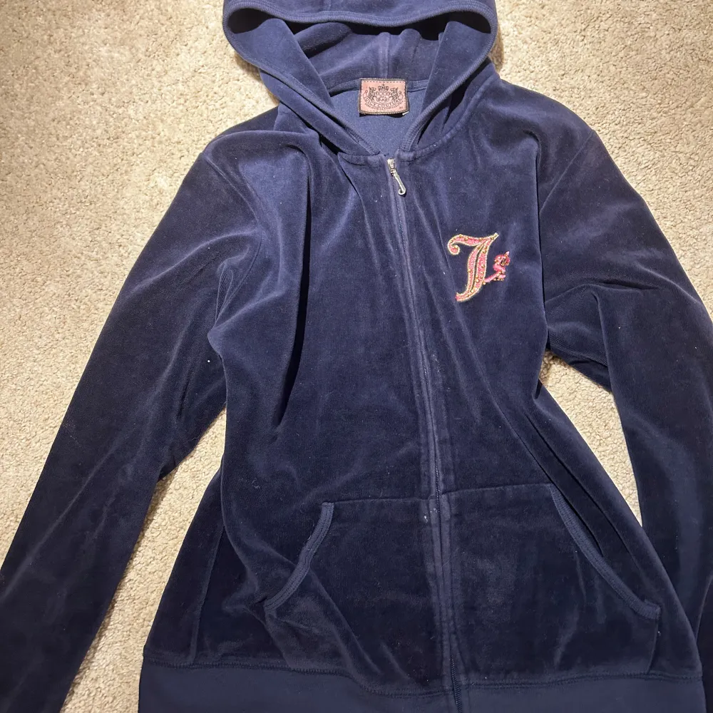 Snygg mörkblå velourjacka från Juicy Couture med dragkedja och huva. Jackan har ett stort, detaljerat broderi på ryggen med guld- och rosa detaljer samt ett litet 'J' på framsidan. Perfekt för en avslappnad och stilren look.. Takit.