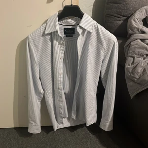 Randig skjorta från Bondelid - Snygg randig skjorta från Bondelid i blått och vitt. Skjortan har en klassisk krage och knappar framtill. Perfekt för en stilren look. Helt ny använd endast en gång.