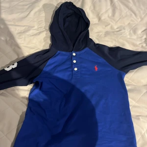 Blå hoodie från Polo Ralph Lauren - Säljer en snygg blå hoodie från Polo Ralph Lauren med mörkblå ärmar och en vit siffra '3' på ena ärmen. Tröjan har en knappslå med tre knappar och en klassisk huva. Perfekt för en avslappnad stil. Pris kan diskuteras!!!