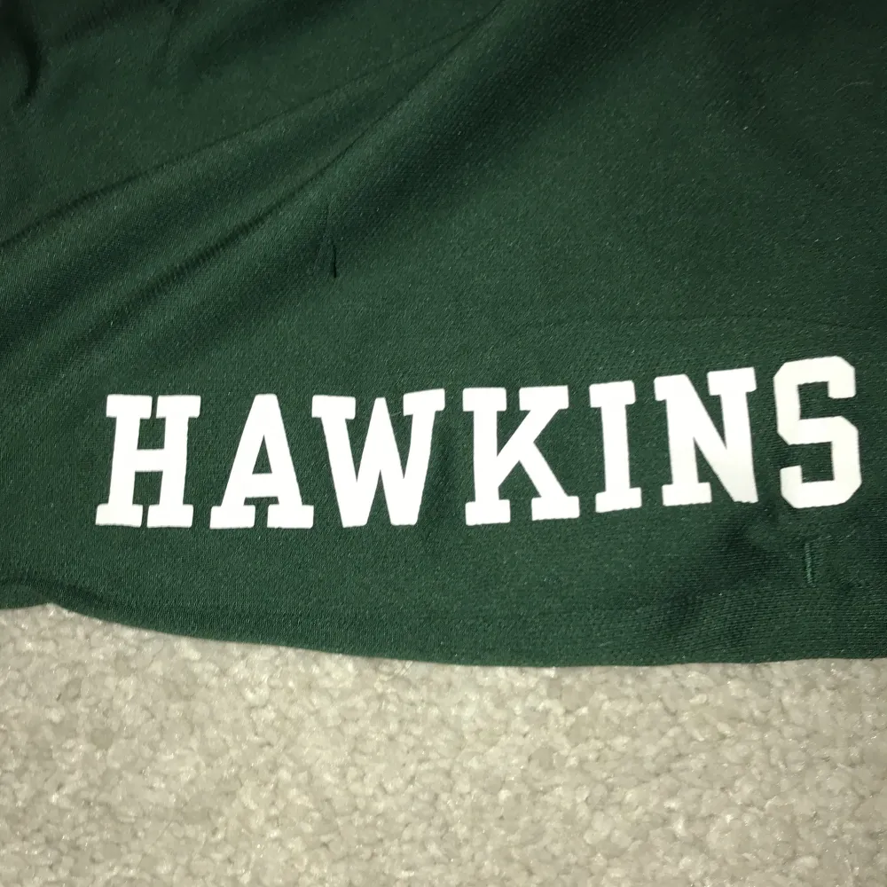 Snygga gröna shorts med texten 'HAWKINS' tryckt i vitt. De har en sportig look med vita ränder vid bensluten. Perfekta för en avslappnad stil eller för att visa din kärlek till serien. Elastisk midja för bekväm passform.de är från basketlaget i stranger things. Sportswear.