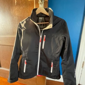 Svart vindjacka från Helly Hansen - Säljer en svart vindjacka från Helly Hansen med Helly Tech-skydd. Jackan har dragkedjor i kontrasterande vitt och rött, samt flera praktiska fickor. Perfekt för blåsiga dagar och utomhusaktiviteter. Kan sälja den för 300 om nån köper snabbt 