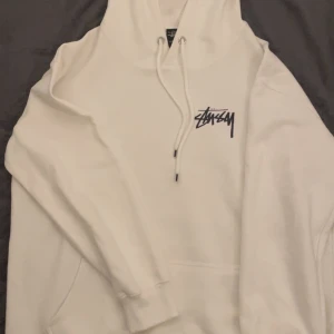 Vit hoodie från Stussy - Säljer en snygg vit hoodie från Stussy med tryck på bröstet och en stor grafisk design på ryggen. Hoodien har en klassisk passform med justerbar dragsko i huvan och en stor magficka. Perfekt för en avslappnad stil! Passar M och S. Använts 1 gång. 