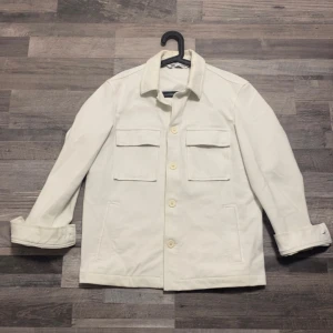 Vit overshirt från Zara - Säljer en stilren vit overshirt från Zara med knappar framtill och två bröstfickor. Jackan har en klassisk krage och långa ärmar. Perfekt för en minimalistisk look. Inga defekter på jackan som jag har märkt. Levererar samma dag som beställningen. Priset kan diskuteras vid snabb och smidig affär.