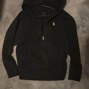 Svart hoodie från Polo Ralph Lauren - Säljer en stilren svart hoodie från Polo Ralph Lauren med dragkedja och det klassiska logotypbroderiet på bröstet. Tröjan har långa ärmar och en praktisk huva med dragsko. Perfekt för en avslappnad look. Stoleken är M men passar bra på S