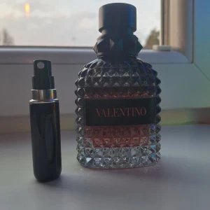 5ml av coral fantasy Valentino parfym - Elegant parfymflaska från Valentino med en lyxig design. Flaskan har en unik, facetterad yta och en mörk etikett med märkesnamnet. Parfymen har en djup och sofistikerad doft, perfekt för speciella tillfällen. Den medföljande svarta sprayflaskan är praktisk för resor.