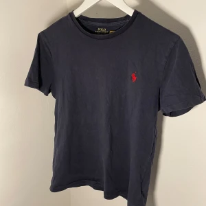 Mörkblå t-shirt från Ralph Lauren - Säljer en stilren mörkblå t-shirt från Ralph Lauren med det klassiska röda broderade logotypen på bröstet. T-shirten har en normal passform och är tillverkad i mjuk bomull.