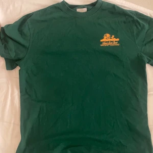 Grön t-shirt från Aimé Leon Dore - Säljer en grön t-shirt från Aimé Leon Dore med tryck i gult på både fram- och baksidan. T-shirten har en klassisk rund hals och korta ärmar. Perfekt för en avslappnad stil. Säljer den för att den är för stor i storleken, köpte den i S men passar någon som har L