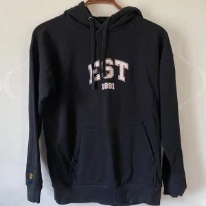 Svart hoodie med EST 1891 tryck - Säljer en svart hoodie med trycket 'EST 1891' på bröstet. Tröjan har en stor ficka framtill och justerbar huva med dragsko. Perfekt för en avslappnad stil. Tröjan är från Lager 157.