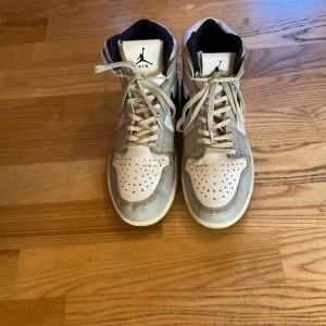 Nike Air Jordans i grått och vitt - Säljer ett par klassiska Nike Air Jordans i grått och vitt. Skorna har en hög modell med snörning och den ikoniska swoosh-loggan i svart på sidan. Perfekta för dig som älskar streetwear och sneakers.
