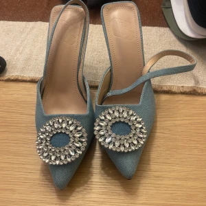 Blå pumps med strassdetalj - Eleganta blå pumps med spetsig tå och en stor glittrig strassdetalj framtill. Skorna har en smal rem runt hälen och är tillverkade i ett jeansliknande material.