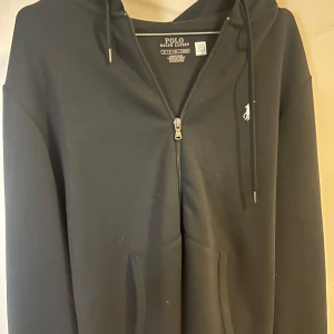 Svart hoodie från Polo Ralph Lauren - Säljer en stilren svart hoodie från Polo Ralph Lauren med dragkedja och klassisk logga på bröstet. Tröjan har en bekväm passform med långa ärmar och en praktisk huva med snörning. Perfekt för en avslappnad look. 9/10 skick.