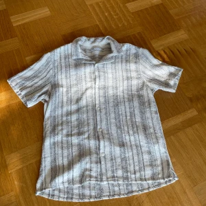 Vit och grå randig skjorta från Zara - Snygg kortärmad skjorta från Zara i vitt med svarta ränder. Skjortan har en avslappnad passform och knappar framtill. Perfekt för varma dagar och ger en stilren look.