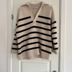 Randig stickad tröja från Zara - Säljer en snygg stickad tröja från Zara i beige och svart med randigt mönster. Tröjan har en v-ringad krage och långa ärmar, perfekt för kyligare dagar. Den har en avslappnad passform och ribbade kanter.