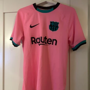 Rosa FC Barcelona fotbollströja från Nike - Säljer en rosa FC Barcelona fotbollströja från Nike med korta ärmar. Tröjan har klubbens emblem och sponsorlogga på framsidan samt Dri-FIT-teknologi för optimal komfort. Perfekt för fans som vill visa sitt stöd med stil! 💖Använd ungefär 2 gånger!