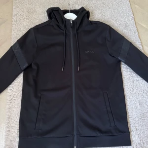 Svart hoodie från Boss - Säljer en stilren svart hoodie från Boss med dragkedja och huva. Jackan har långa ärmar och diskreta fickor framtill. 