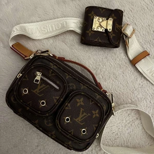 Brun axelväska  - Snygg brun axelväska med klassiskt monogrammönster och flera praktiska fack. Väskan har en justerbar axelrem i vitt med märkets namn och detaljer i guld. Perfekt för att bära med sig det viktigaste med stil. Super fin och elegant dessutom helt oanvänd och ny