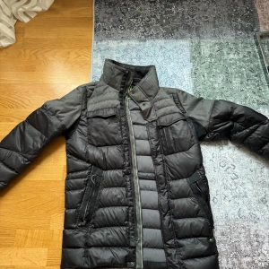 Svart pufferjacka från Boss - Säljer en stilren svart pufferjacka från Boss med quiltad design och hög krage. Jackan har långa ärmar och praktiska dragkedjor på fickorna. Perfekt för kyliga dagar.