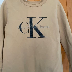 Beige tröja från Calvin Klein Jeans - Säljer en snygg beige tröja från Calvin Klein Jeans med CK-logon tryckt på framsidan. Tröjan har ärmar och en rund halsringning. 