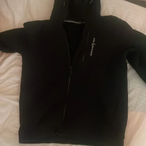 Svart huvtröja från Sail Racing - Säljer en svart huvtröja från Sail Racing med dragkedja framtill. Tröjan har en stilren design med logotyp på bröstet och en bekväm huva. Perfekt för en avslappnad stil.