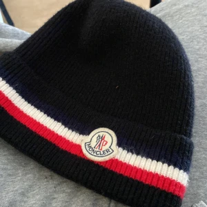 Svart mössa från Moncler - Snygg svart mössa från Moncler med röd och vit randdetalj. Den har en broderad Moncler-logga framtill. Perfekt för att hålla sig varm och stilren under kyliga dagar. Scannen kmr direkt till monclers hemsida
