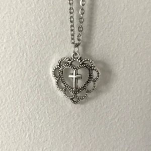 Silverfärgat halsband med hjärta och kors - Elegant halsband i silverfärg med ett detaljerat hjärthänge som har ett kors i mitten. Hänget har en vacker, spetsliknande design som ger en klassisk och tidlös känsla. Perfekt för att ge en subtil men meningsfull touch till din stil.