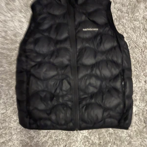 Svart dunväst från Peak Performance - Svart dunväst från Peak Performance med quiltad design och dragkedja framtill. Västen har en stilren look med diskret logga på bröstet och är tillverkad i lätt och smidigt material. Perfekt att slänga över en hoodie eller tröja när det är kyligt ute. Ända är att det är ett igen sytt jack på västen.