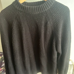 Svart ribbstickad tröja - En svart, ribbstickad tröja med rund hals och långa ärmar. Tröjan har en chunky känsla och bred ribbad mudd vid halsen, perfekt för lager på lager under kyliga dagar. Enkel och stilren design som passar till det mesta.