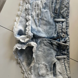 Ljusblå lågmidjade jeansshorts  - Supersnygga ljusblå jeansshorts med slitna detaljer och vit spets längs kanterna. De har låg midja, klassiska fickor och dragkedja framtill. Perfekta för sommarens alla äventyr och ger en cool, avslappnad vibe.