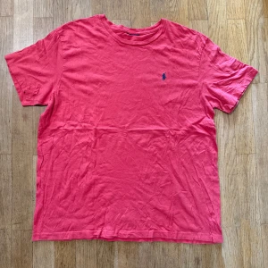 Rosa t-shirt från Ralph Lauren - Snygg rosa t-shirt från Ralph Lauren med klassisk blå broderad logga på bröstet. T-shirten har rund halsringning och korta ärmar. Tillverkad i mjuk bomull som känns skön mot huden. Perfekt för en chill och stilren look.