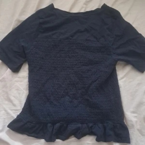 Mörkblå topp med volang från Gina Tricot - Supersöt mörkblå topp från Gina Tricot med smockad framsida och volangkant nertill. Toppen har korta ärmar och en mjuk, stretchig känsla. Perfekt för dig som gillar en enkel men ändå söt stil.