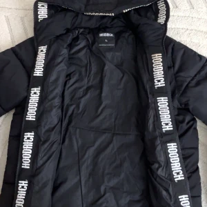 Svart pufferjacka från Hoodrich L - Svart pufferjacka från Hoodrich med logga tryckt på bröstet och längs dragkedjan. Jackan har huva, raka sömmar och två sidofickor. Perfekt för kalla dagar och har en clean streetstyle-vibe.